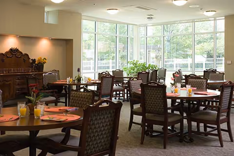 A bright dining room with multiple round tables set with orange placemats, glasses of orange juice, and silverware. The room has large windows allowing natural light to fill the space, and there are several upholstered chairs around each table. A wooden sideboard with decorative items and flowers is visible against one wall.