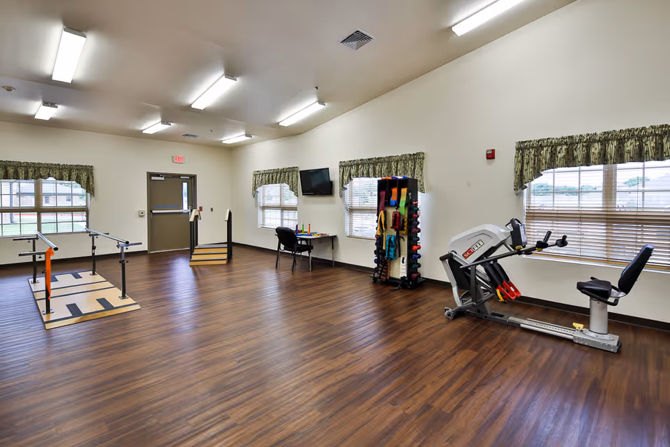A spacious senior rehabilitation room with parallel bars, exercise equipment, a rack of weights and bands, chairs, and large windows.