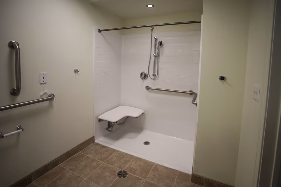 A walk-in shower with white tiled walls and a fold-down seat. The shower has a handheld showerhead and multiple stainless steel grab bars for support. The floor outside the shower is tiled in a brown pattern, and the walls are painted light beige.