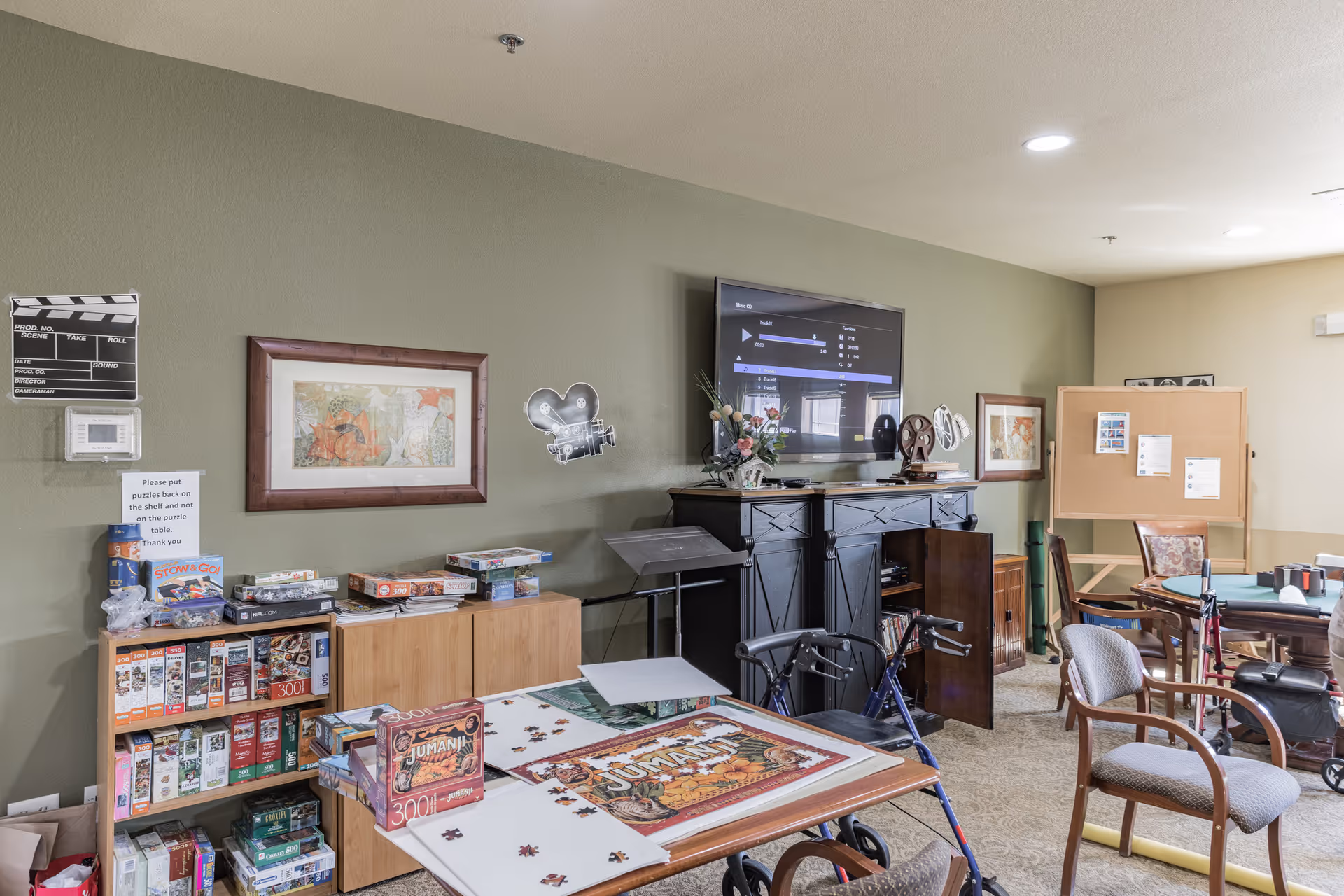 Community activity room with shelves of board games, a table holding a partially completed puzzle, a TV on a cabinet, and chairs and walkers.