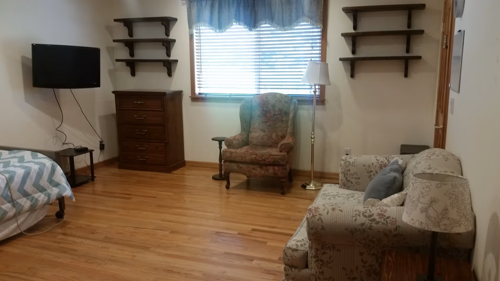 A cozy room with wooden flooring featuring a bed with a chevron patterned blanket on the left, a wall-mounted flat screen TV above a small table, a wooden chest of drawers, a floral upholstered armchair near a window with blinds and a blue valance, a standing lamp, and a floral patterned sofa with cushions and a matching lamp on a side table.
