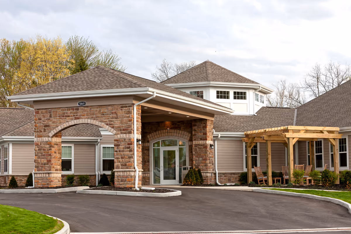 Exterior view of Pinebrook of Milford by StoryPoint Group, showing the entrance with a covered driveway supported by stone pillars, a wooden pergola with chairs on the right, and a building with multiple windows and a shingled roof surrounded by trees.