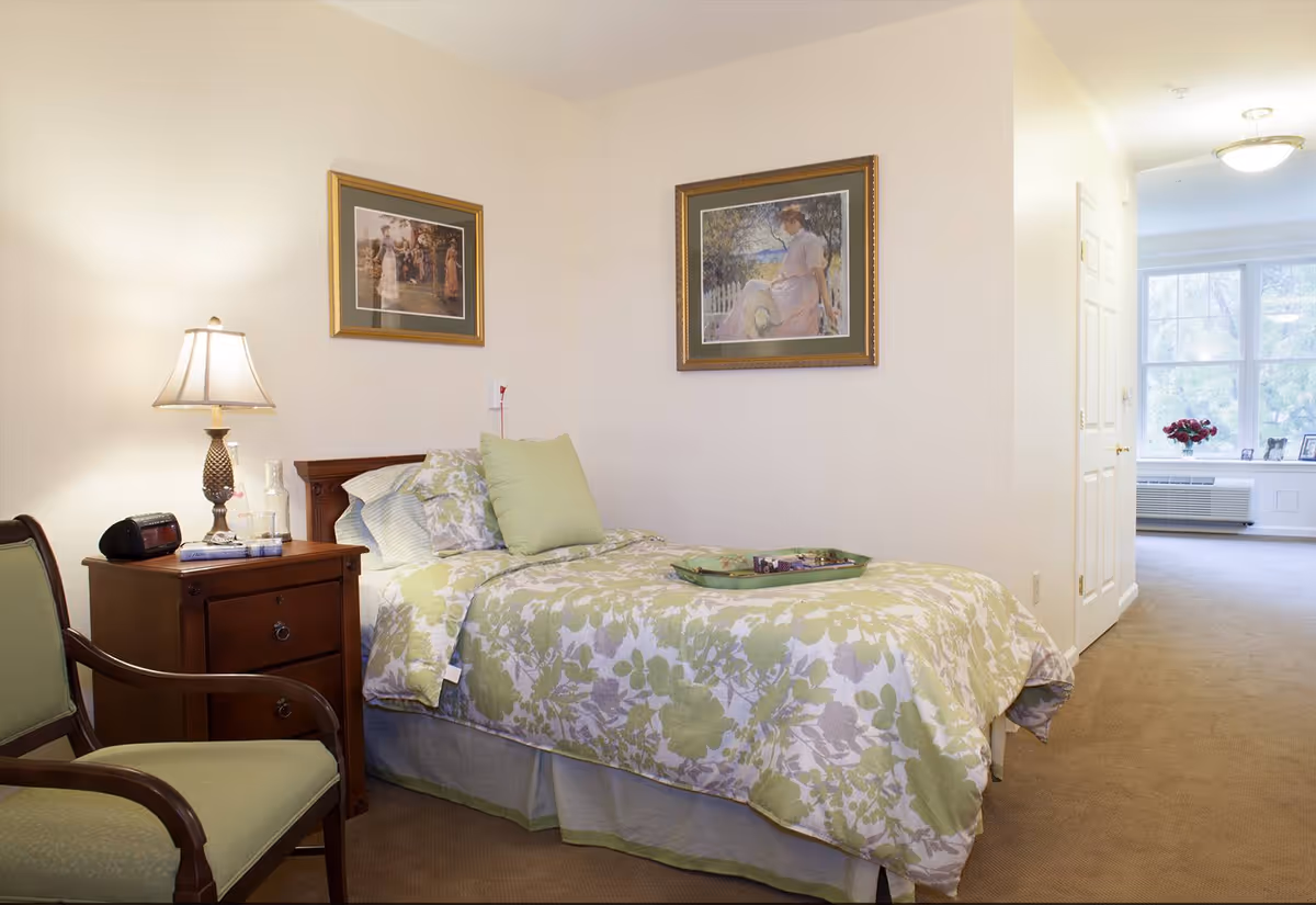 A cozy bedroom with a single bed covered in a green and white floral bedspread. There are two framed paintings on the walls above the bed. Next to the bed is a wooden nightstand with a lamp, a clock radio, and some small items. A green cushioned wooden chair is positioned beside the nightstand. The room has beige carpet and light-colored walls, with a hallway leading to a bright windowed area in the background.