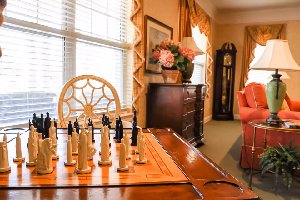 A cozy living room area with a wooden chessboard and chess pieces in the foreground. Behind the chessboard, there is a decorative chair, a wooden dresser with a flower pot on top, a tall grandfather clock, a red armchair, and a side table with a green lamp. The room has large windows with blinds and patterned curtains.