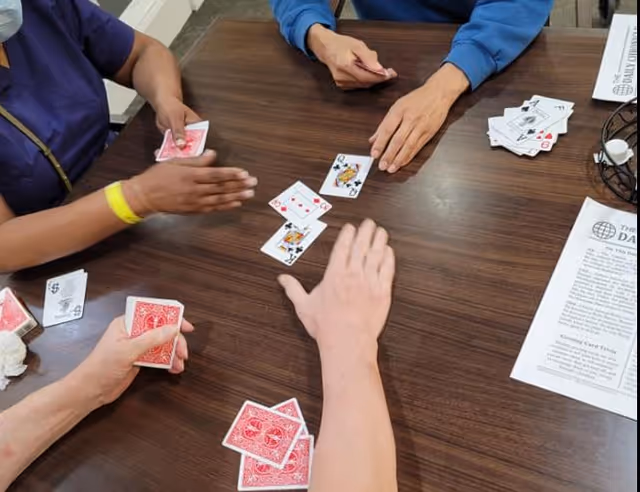 Four people sitting around a wooden table playing a card game. Several playing cards are visible on the table, along with a printed sheet of paper and a wire basket.