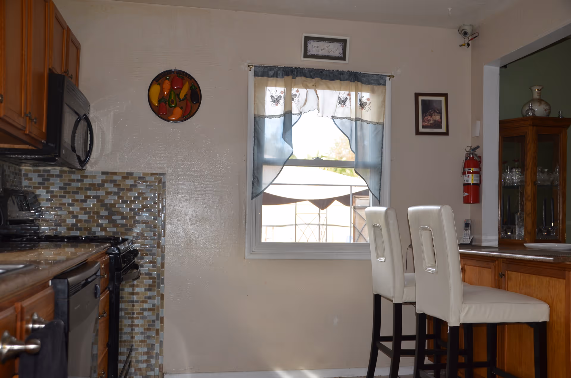 Interior view of a kitchen with wooden cabinets, a tiled backsplash, a stove, microwave, and dishwasher on the left side. On the right side, there is a counter with two white cushioned bar stools. A window with blue and white curtains is centered on the wall, and a fire extinguisher is mounted on the wall near a glass cabinet with decorative items.