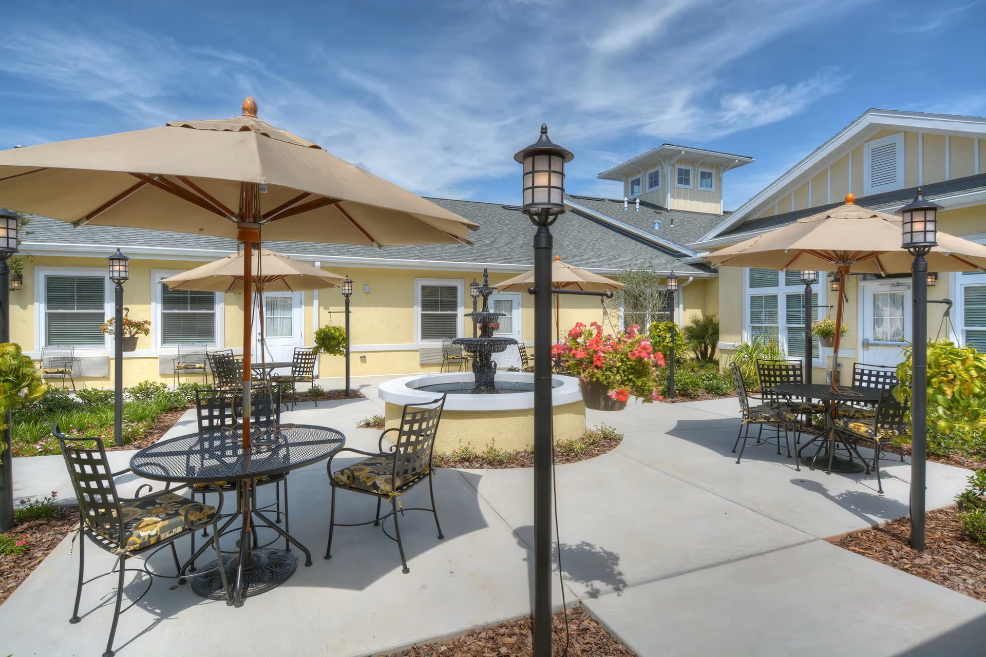 Outdoor courtyard area at Benton House of Clermont featuring several black metal tables with patterned cushions and large beige umbrellas. There is a central water fountain surrounded by a circular planter. The courtyard is paved with concrete walkways and decorated with hanging flower baskets and garden plants. The building exterior is painted yellow with white trim under a blue sky with some clouds.