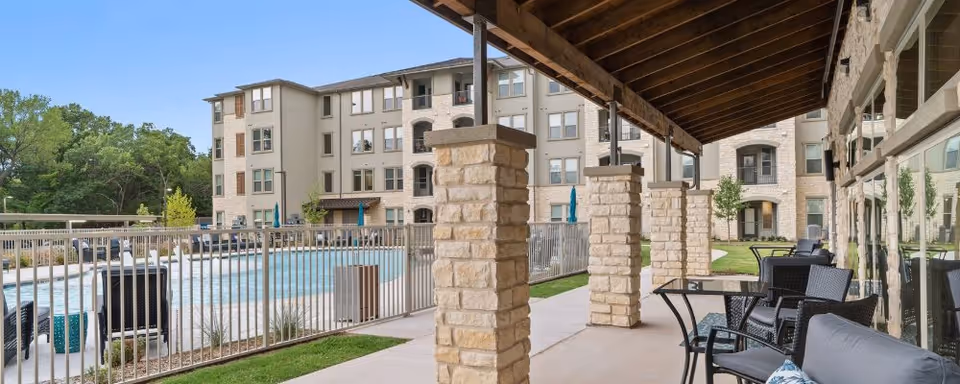 Covered patio with chairs and tables overlooking a fenced outdoor swimming pool in front of the apartment building.