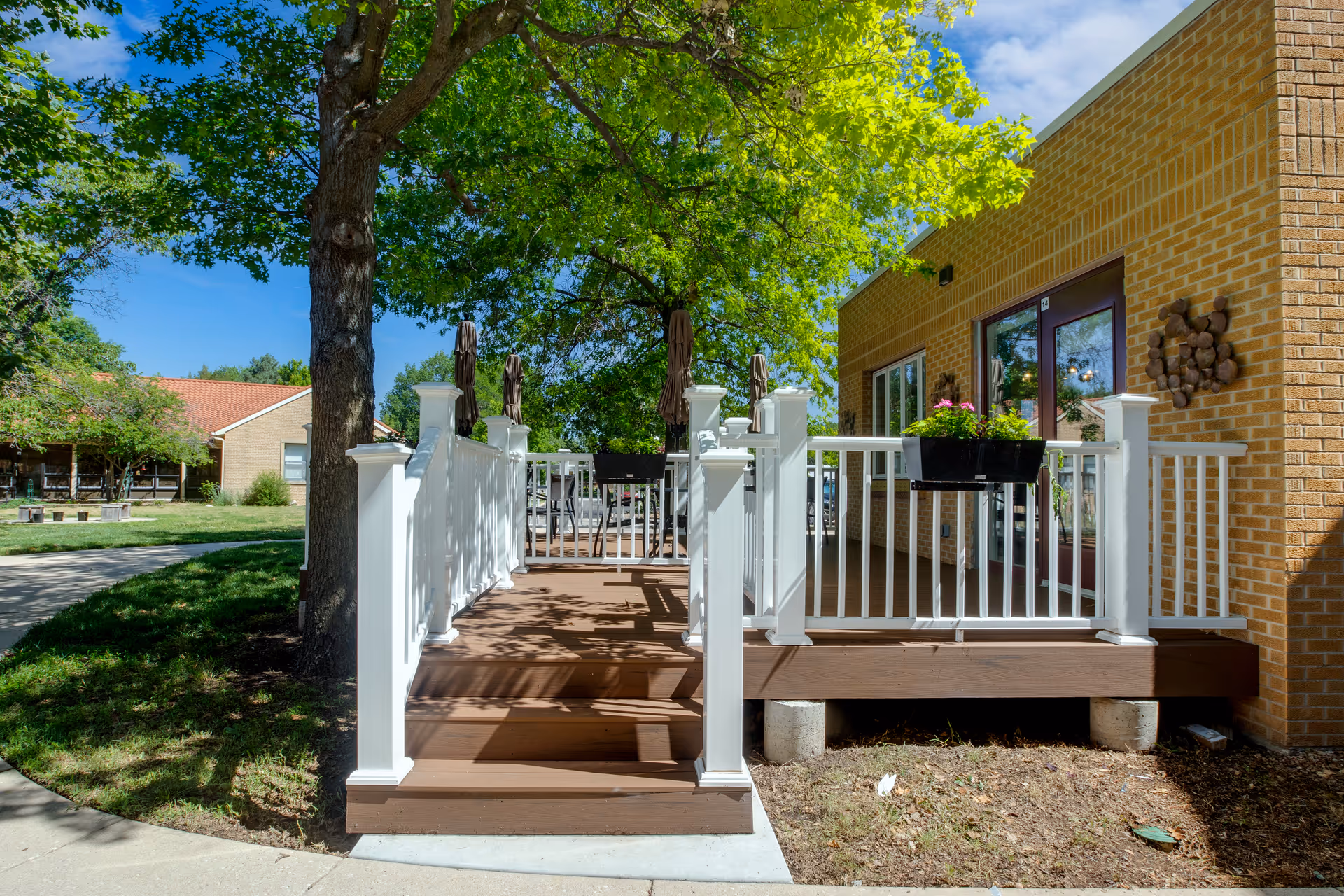 Outdoor patio area with a wooden deck and white railing attached to a brick building. There are potted flowers on the railing and several closed umbrellas on the deck. A large tree provides shade, and there is a grassy area and sidewalk nearby under a clear blue sky.