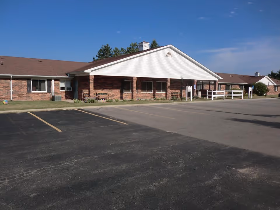 Exterior view of a single-story brick building with a white gabled entrance and a brown roof under a clear blue sky. There is a paved parking lot in front with marked parking spaces and some benches near the entrance.