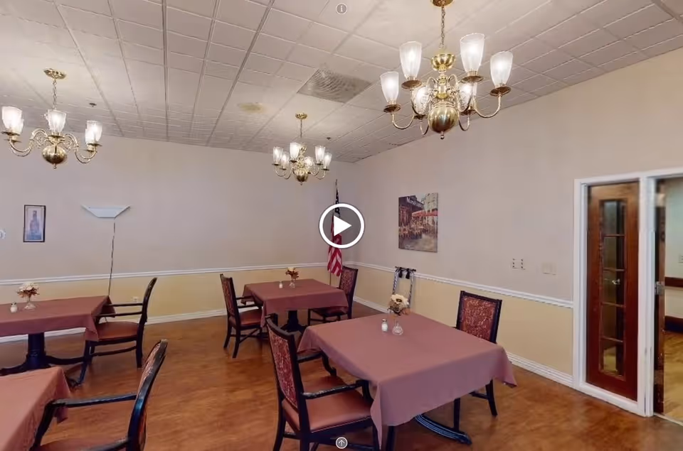 A dining room with several tables covered in maroon tablecloths, each surrounded by chairs with patterned upholstery. The room has wooden flooring, beige walls with a white chair rail, and three brass chandeliers with frosted glass shades hanging from a tiled ceiling. There is an American flag in the corner, a floor lamp, framed artwork on the walls, and a doorway with glass-paneled double doors leading to another room.
