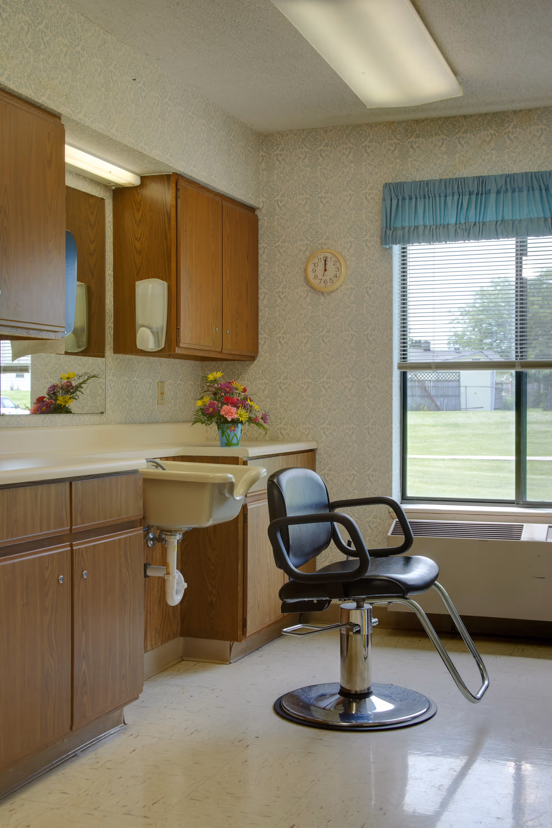 Interior grooming area with a black salon chair in front of a sink and wooden cabinets beside a window.