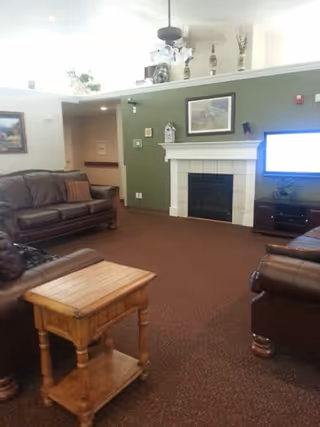 A cozy living room with brown leather sofas arranged around a wooden side table. The room features a green accent wall with a white fireplace and a mounted flat-screen TV. Decorative items and framed artwork are displayed above the fireplace and on the walls.