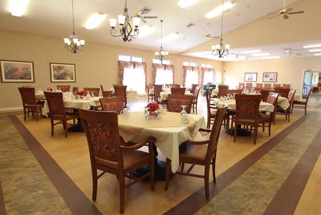 Spacious dining room with multiple round tables set with tablecloths and chairs under chandeliers and large windows.