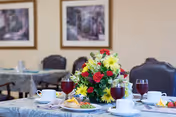 A dining table set with plates of food, cups, and glasses of red beverage. A centerpiece of colorful flowers including red, yellow, and white blooms is placed in the middle of the table. In the background, there are additional tables and chairs, and framed artwork on the wall.