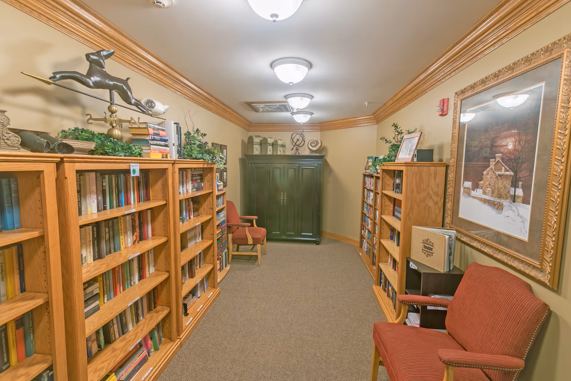 A cozy library room with wooden bookshelves filled with books on both sides. There are two red upholstered chairs, one on each side of the room. The walls are painted beige with wooden crown molding. A large framed painting hangs on the right wall above one of the chairs. The ceiling has three light fixtures, and there is a dark green cabinet at the far end of the room with decorative items on top.