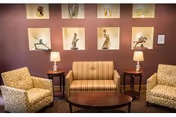 A cozy sitting area with a striped loveseat flanked by two patterned armchairs. Two wooden side tables each hold a lamp with a beige shade. The wall behind the seating features nine illuminated display niches showcasing various small sculptures and art pieces.