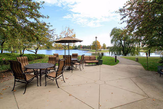 Outdoor patio area with metal tables and cushioned chairs, one table with a large umbrella, surrounded by trees and overlooking a calm lake with a walking path nearby under a partly cloudy sky.