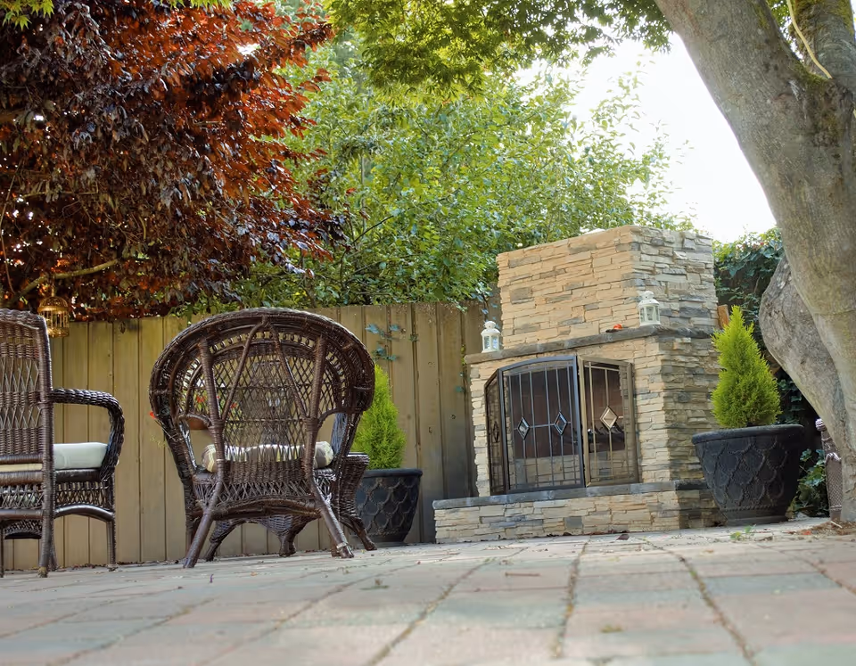 Outdoor patio area with wicker chairs and a stone fireplace surrounded by potted plants and trees, enclosed by a wooden fence.