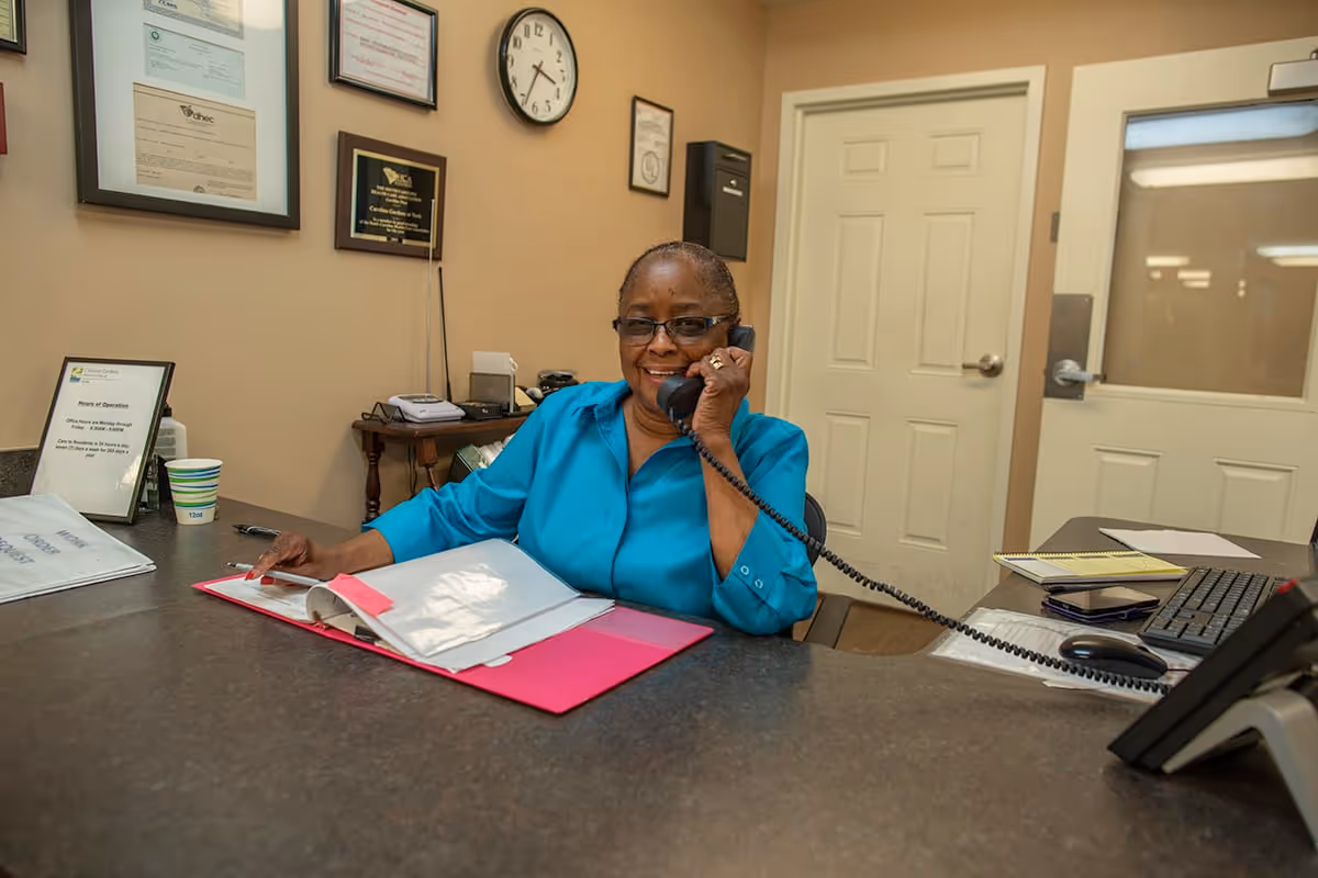 A woman wearing glasses and a blue shirt is sitting at a reception desk, talking on a corded telephone. The desk has an open binder with papers, a keyboard, a mouse, a notepad, and a cup. Behind her are framed certificates on the wall, a clock showing 10:10, and a door with a window.