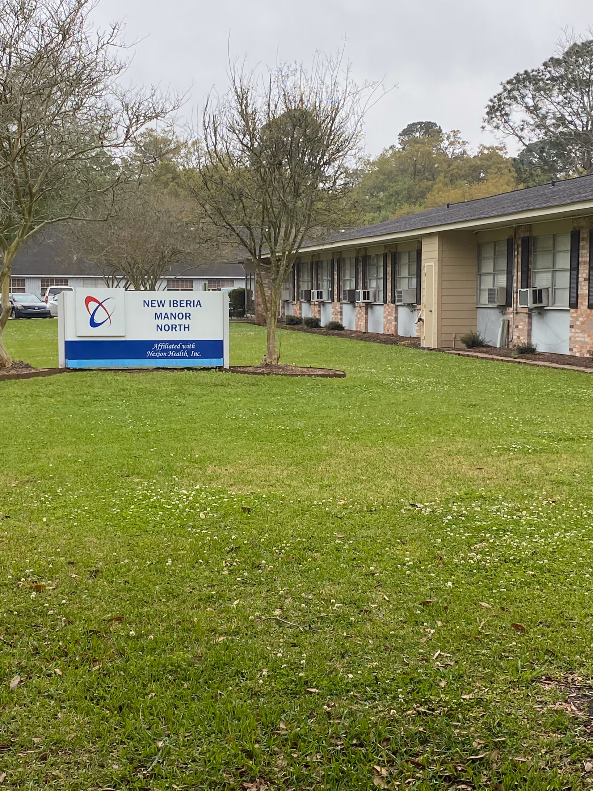 Exterior view of New Iberia Manor North facility showing a green lawn with trees and a sign in front that reads 'New Iberia Manor North Affiliated with Nexon Health, Inc.' The building has multiple windows with air conditioning units and a brick and siding exterior.