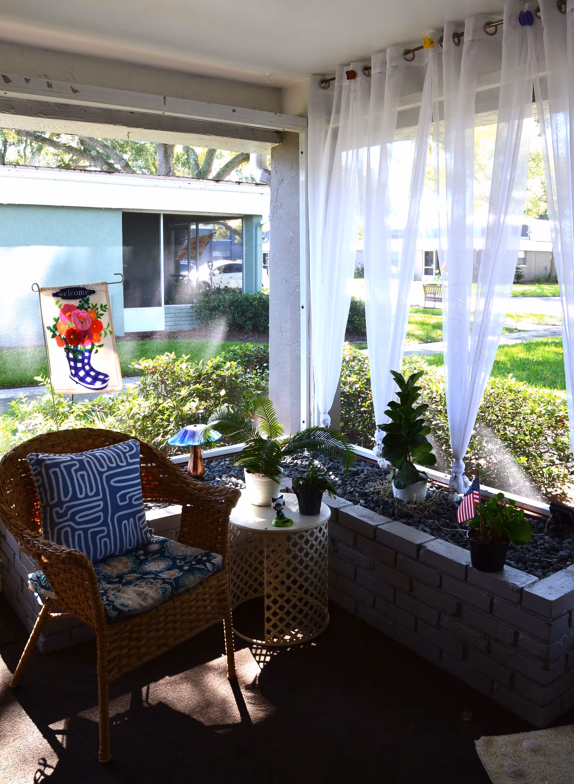 A cozy screened-in porch area with a wicker chair featuring patterned cushions, a small round white table with potted plants, and sheer white curtains hanging from the ceiling. Outside the porch, there is a garden with green grass, bushes, and a small American flag in a plant pot. A colorful welcome flag with flowers is also visible outside.