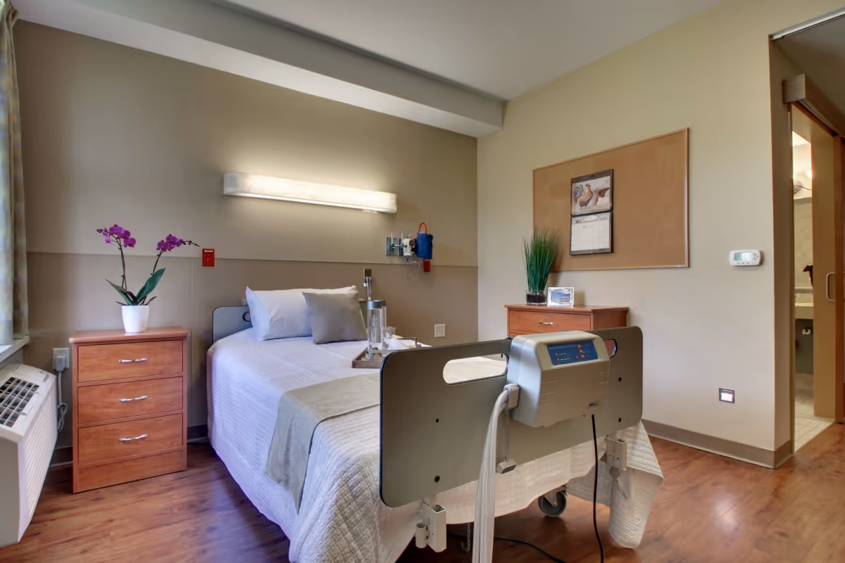 A neatly made single hospital-style bed with white and beige bedding in a senior living facility room. The bed has medical equipment attached to the footboard. There are two wooden nightstands, one with a potted orchid and the other with a green plant and a framed photo. A calendar is pinned to a corkboard on the wall. The room has wood flooring and a window with curtains on the left side. A bathroom entrance is visible on the right.