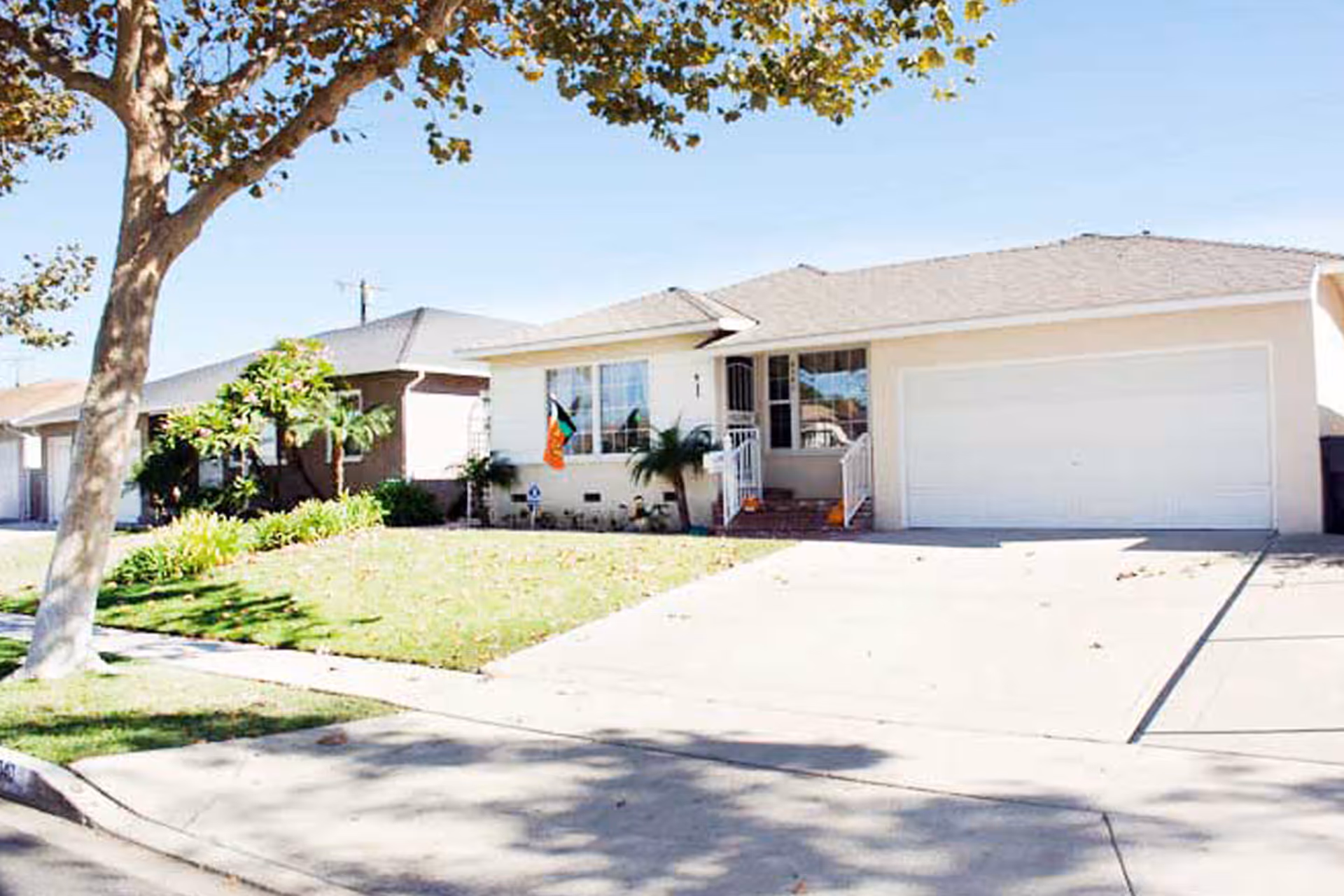 Single-story residential house with a light-colored exterior, a two-car garage, a small front porch with steps, and a well-maintained lawn with a tree and some plants. The sky is clear and blue.
