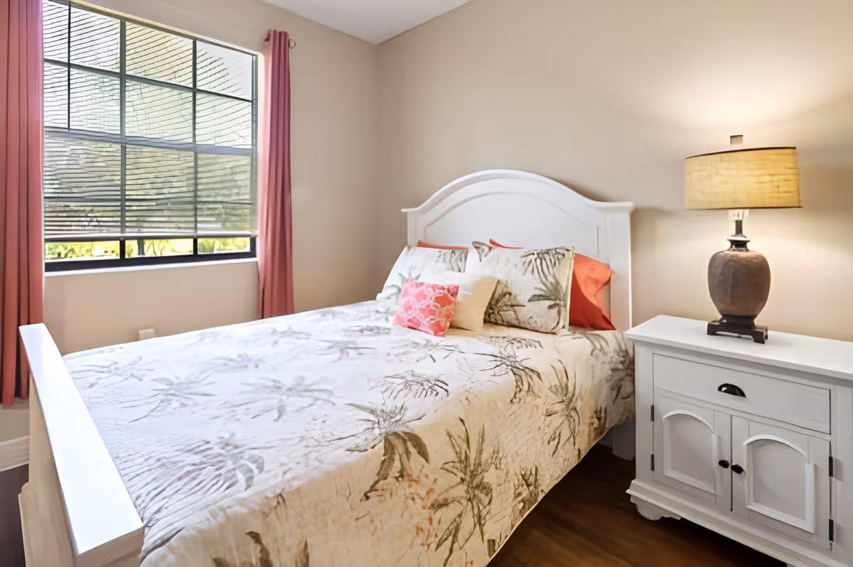 A bright bedroom with a white wooden bed frame and a bed covered with a floral patterned quilt and multiple pillows. Next to the bed is a white nightstand with a brown table lamp. A window with pink curtains and partially closed blinds lets in natural light.