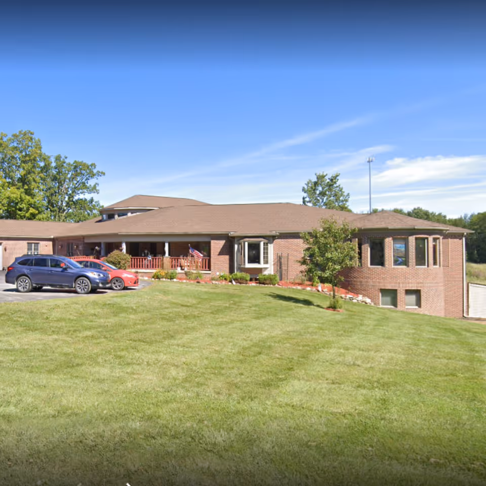 Exterior view of Courtyard Manor of Howell, a single-story brick building with a brown roof, surrounded by a well-maintained grassy lawn. There are two cars parked in front of the building, and a small tree and shrubs are visible near the entrance. The sky is clear and blue.