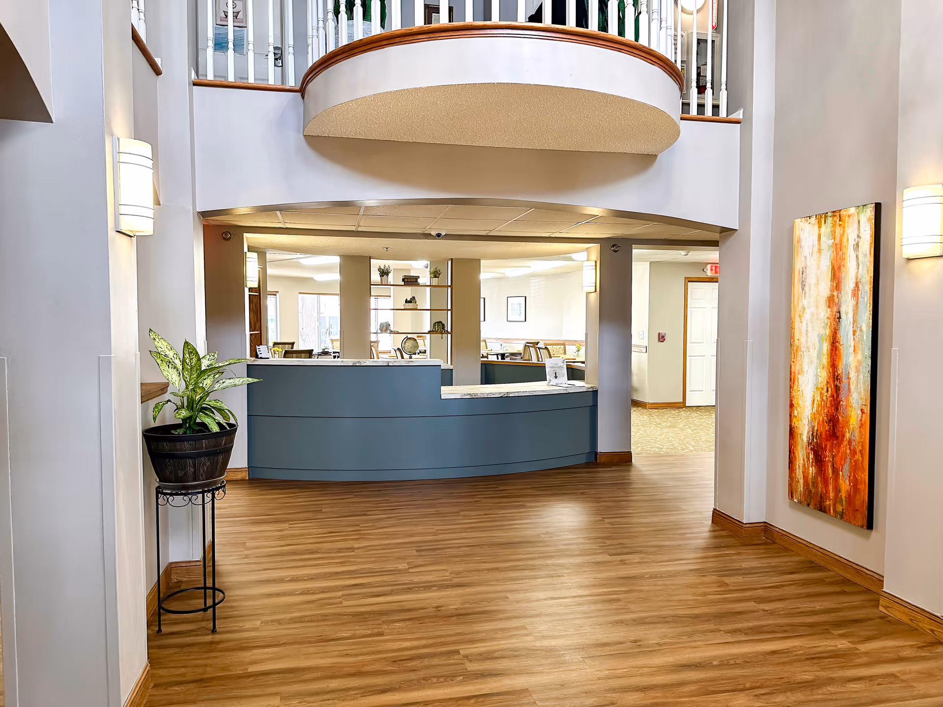 Open reception lobby with a curved blue front desk, wood floors, potted plant, and abstract wall art under a second-floor balcony.
