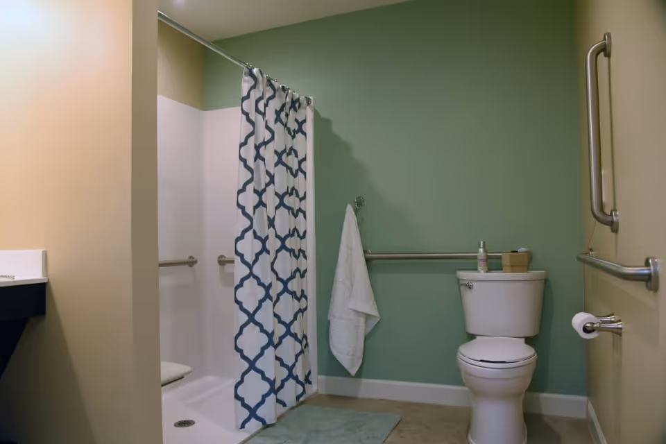 A bathroom with a white toilet, a towel hanging on a grab bar, a shower with a white and blue patterned curtain, and beige and green walls. There are additional grab bars near the toilet and shower for accessibility.