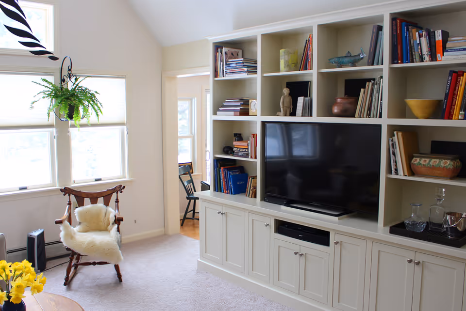 A bright living room with a built-in white shelving unit holding books, decorative items, and a flat-screen TV. A wooden chair with a white fur throw is positioned near two windows with blinds. A hanging green plant is visible near the windows, and a small table with yellow flowers is partially visible in the foreground.