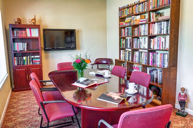 A communal reading/meeting room with a polished oval table surrounded by red chairs, bookshelves, a wall-mounted TV, and a vase of flowers on the table.