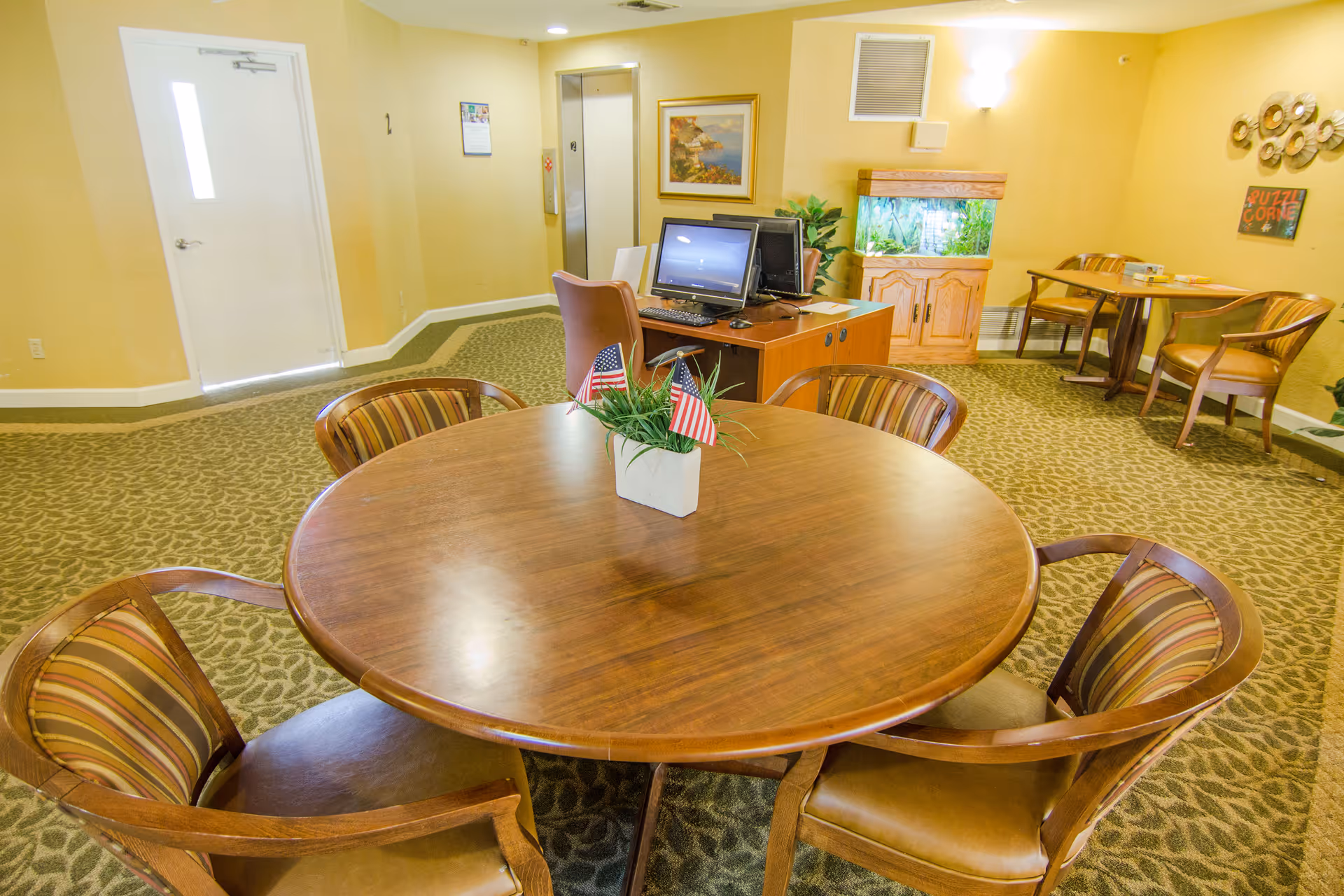 Interior room with a round wooden table surrounded by six striped cushioned chairs. On the table is a small plant with two American flags. In the background, there is a desk with two computer monitors and a chair, a fish tank on a wooden stand, and another small table with two chairs. The walls are painted yellow and decorated with a framed picture and wall art. There is a door and an elevator visible on the left side.