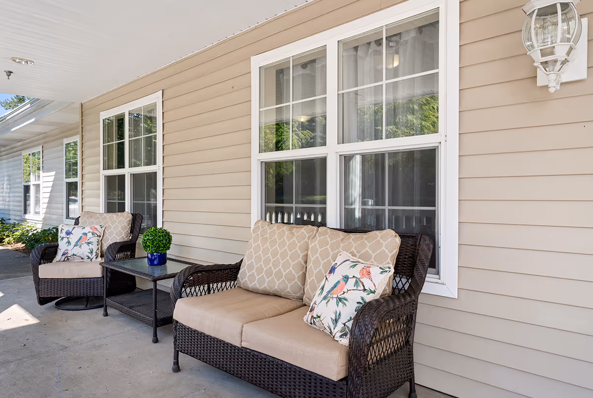 Outdoor seating area on a covered porch with two cushioned wicker chairs and a matching loveseat, each with decorative pillows. A small table with a potted plant sits between the chairs. The porch is attached to a beige siding building with white-framed windows and a wall-mounted lantern light.