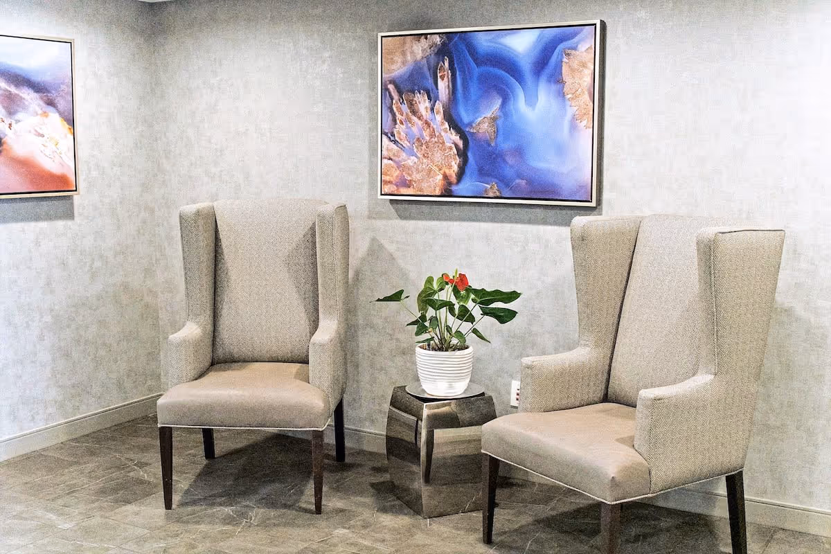 Two beige upholstered wingback chairs positioned on either side of a small metallic side table with a white potted plant featuring green leaves and red flowers. The background wall is light gray with two framed abstract artworks hanging above the chairs.