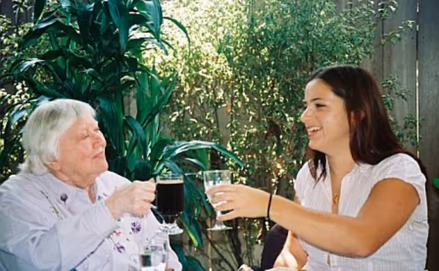 An elderly woman and a younger woman sitting together outdoors, smiling and clinking their glasses in a toast. There are green plants and a fence in the background.