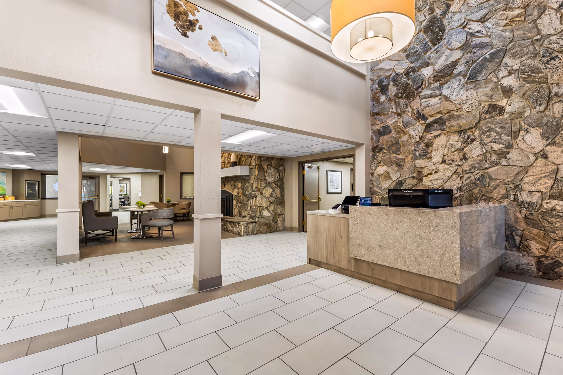 Interior view of a senior living facility lobby area featuring a reception desk with a printer, a stone accent wall, a large hanging light fixture, and a seating area with tables and chairs in the background. The floor is tiled, and there is a painting hanging above the entrance to the seating area.