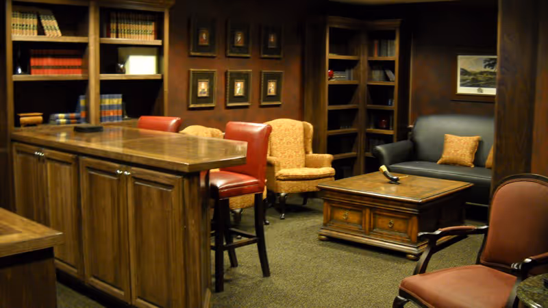 A cozy interior room with dark wood furniture including a tall counter with two red bar stools, a wooden coffee table, a black leather couch with an orange pillow, two patterned armchairs, and bookshelves filled with books. The walls are decorated with framed pictures.