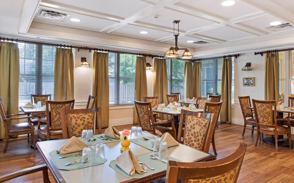 Spacious dining room with multiple set tables and patterned wooden chairs, large windows with curtains, and overhead pendant lighting.