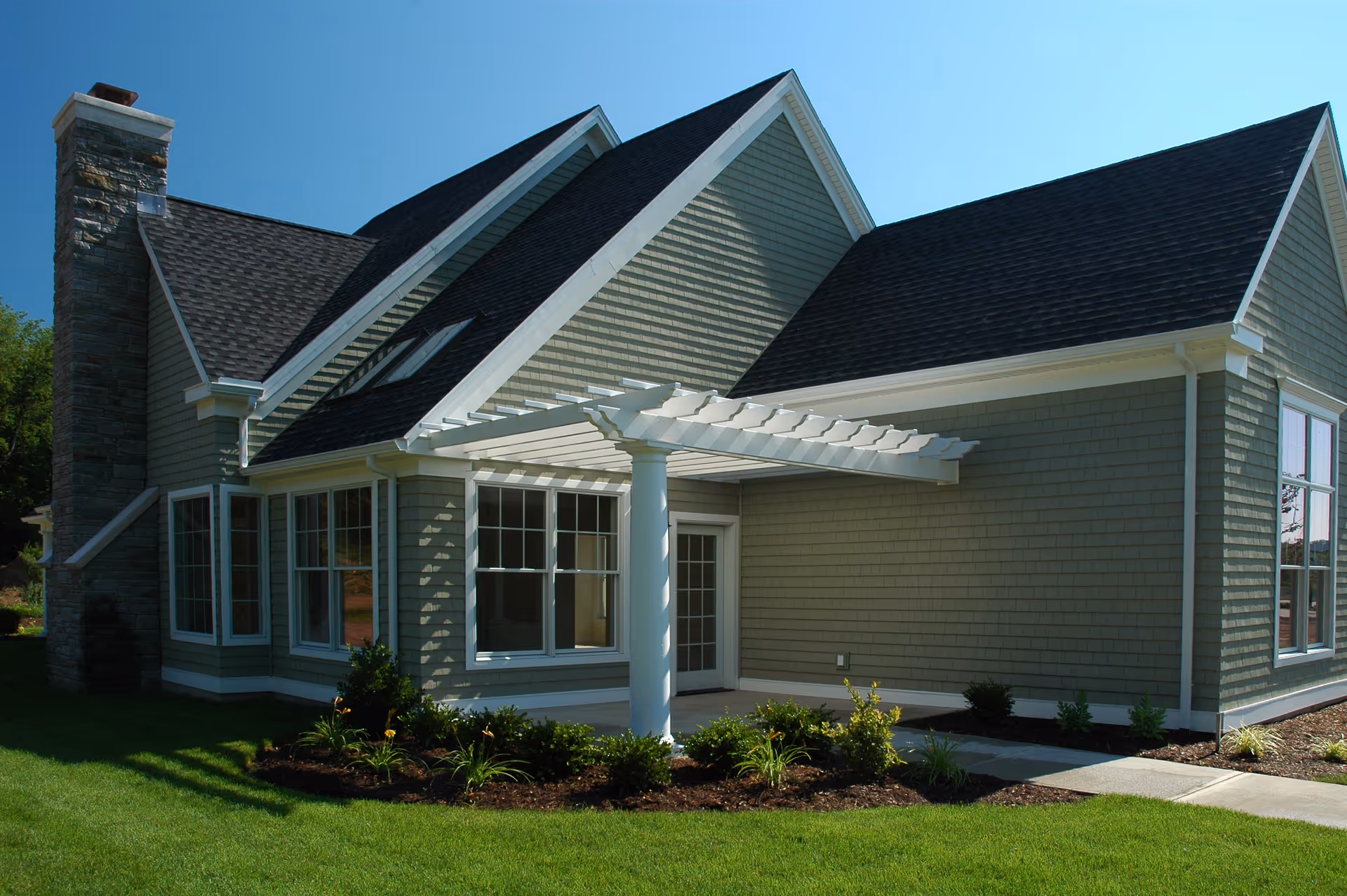 Exterior view of a residential-style building with gray siding, white trim, and a dark shingled roof. The building features a stone chimney, multiple windows, and a white pergola over a small patio area. There is a well-maintained lawn and landscaped garden beds surrounding the building.