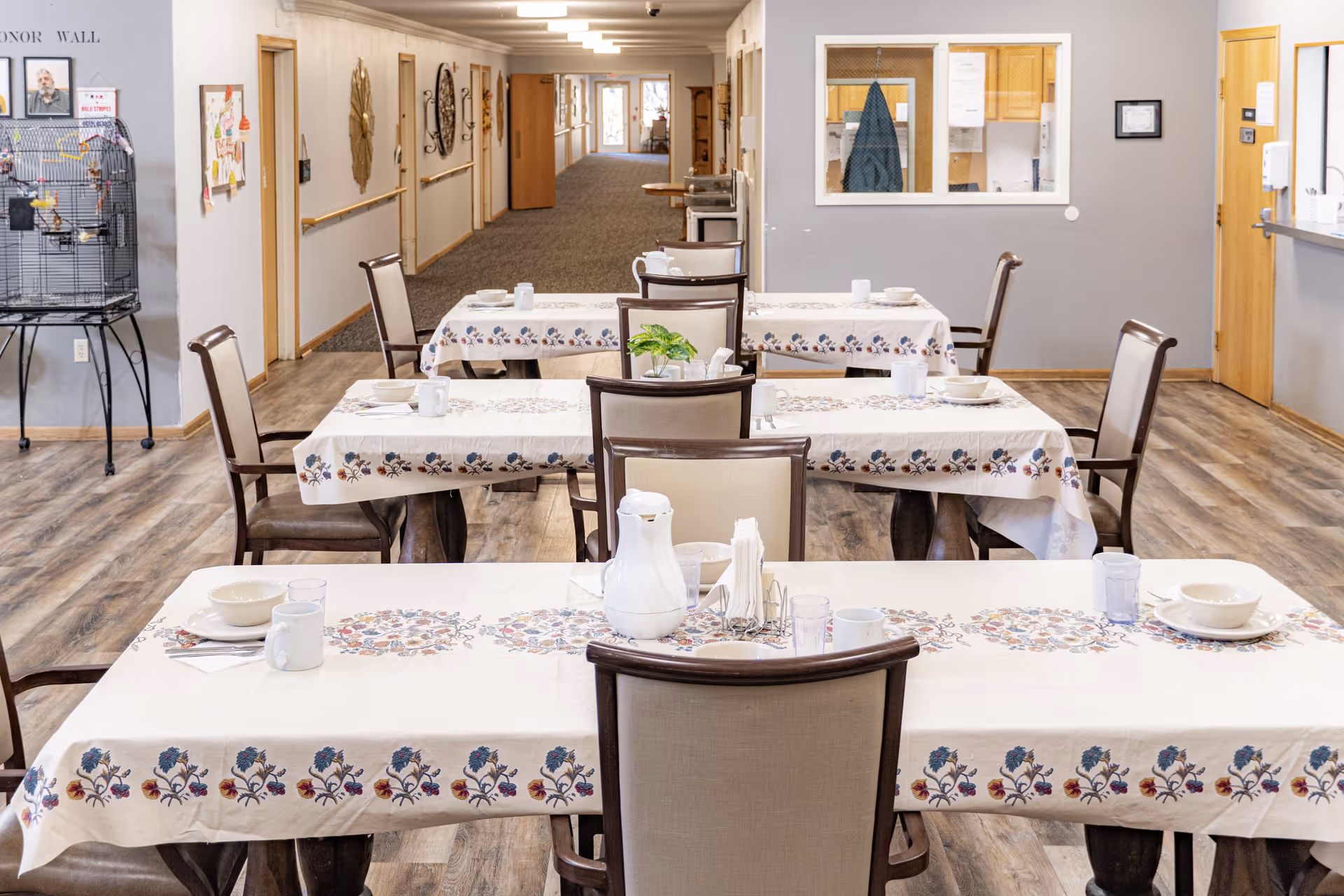 A dining area in Southern Oaks Place with three tables covered in white tablecloths featuring floral patterns. Each table is set with bowls, cups, glasses, and a white pitcher. There are cushioned chairs around the tables. The background shows a long hallway with doors and wall decorations, a birdcage with a bird on the left, and a window into an office area on the right.