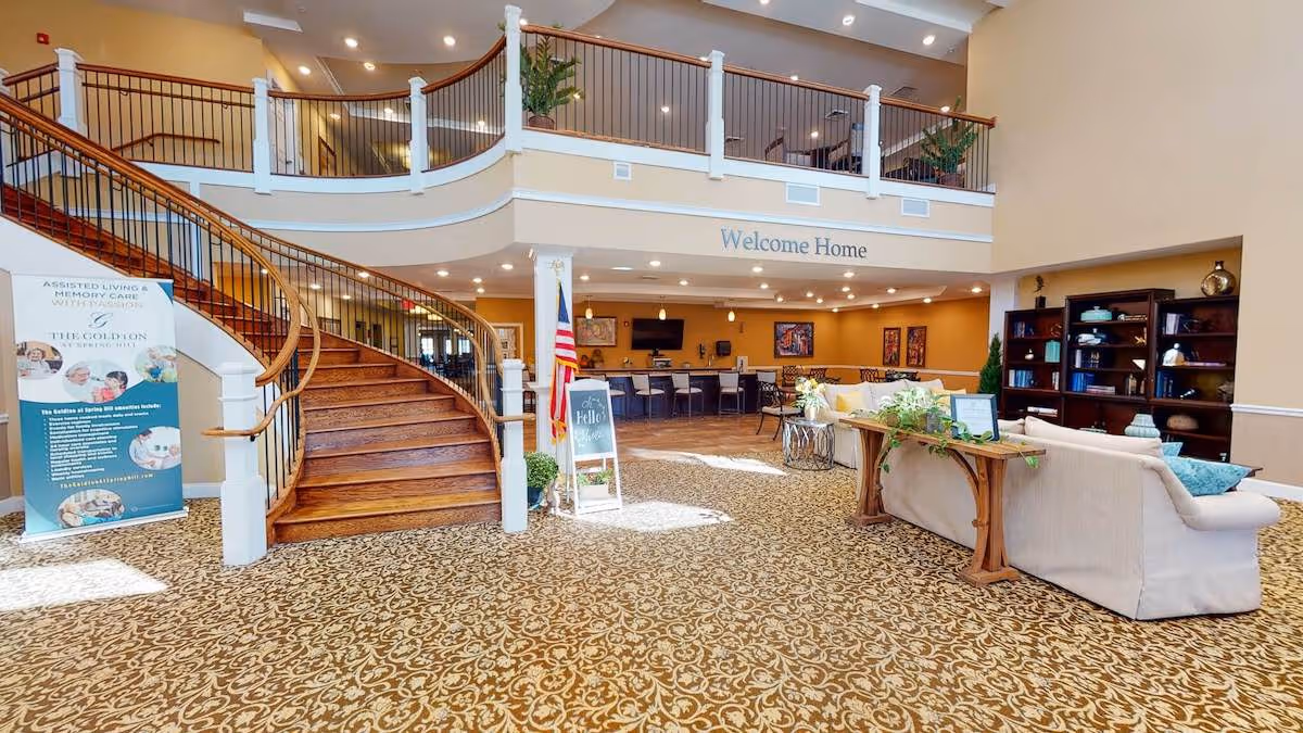 Spacious senior living facility lobby with a curved wooden staircase, patterned carpet, seating area with sofas and a bookshelf, and a sign that reads 'Welcome Home'. There is also an American flag and a banner with information about assisted living and memory care services.