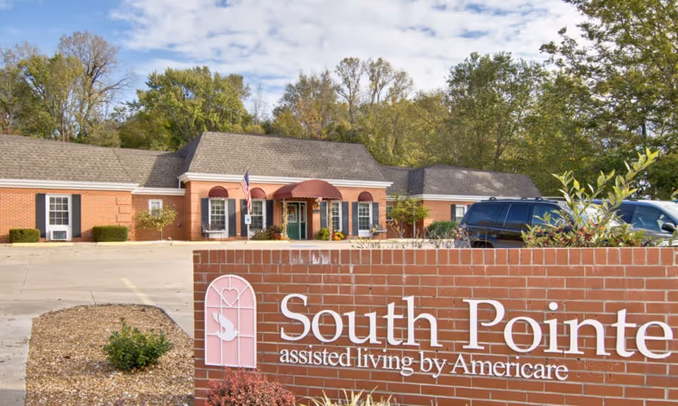 Exterior view of South Pointe assisted living facility showing a brick building with a peaked roof, several windows with black shutters, an entrance with a red awning, and a parking lot with vehicles. In the foreground, there is a brick sign with the facility name and logo.