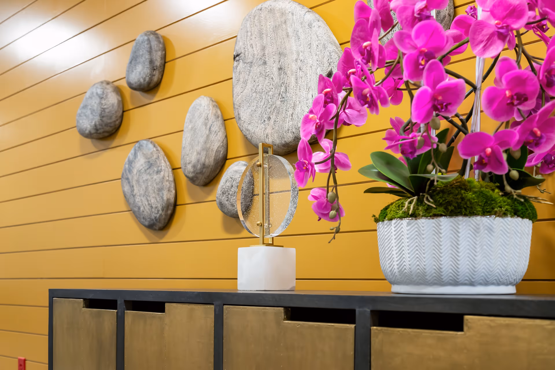 Decorative interior scene featuring a mustard yellow shiplap wall with six stone-like wall hangings arranged in a cluster. In front of the wall is a black and gold cabinet with a modern decorative sculpture and a white textured pot holding vibrant pink orchids with green moss at the base.