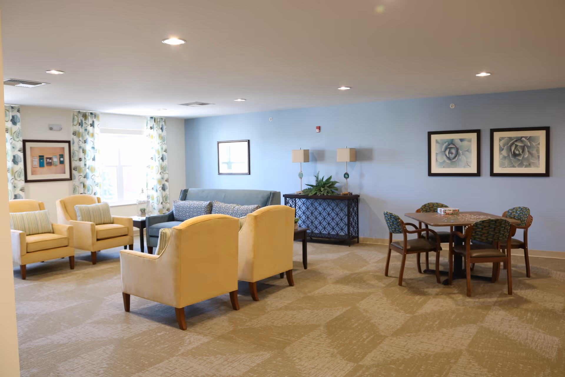 A spacious living room area with beige carpet and light blue walls. The room features a blue sofa with patterned cushions, four beige armchairs with striped pillows, a small black coffee table, and a black console table with two lamps and a plant. There is also a square table with four chairs, a puzzle in progress on the table, and framed artwork on the walls. Light streams in through a window with floral curtains.