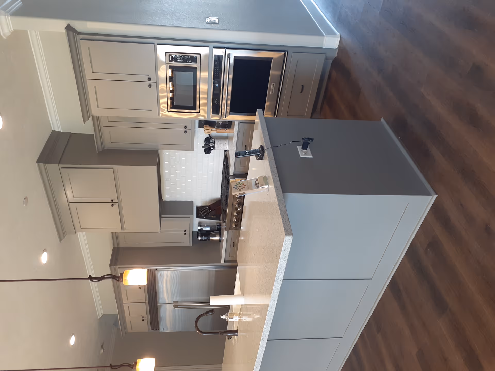 Modern kitchen with gray cabinetry, a large island with a white countertop, stainless steel refrigerator, built-in microwave and oven, pendant lights hanging above the island, and hardwood flooring.