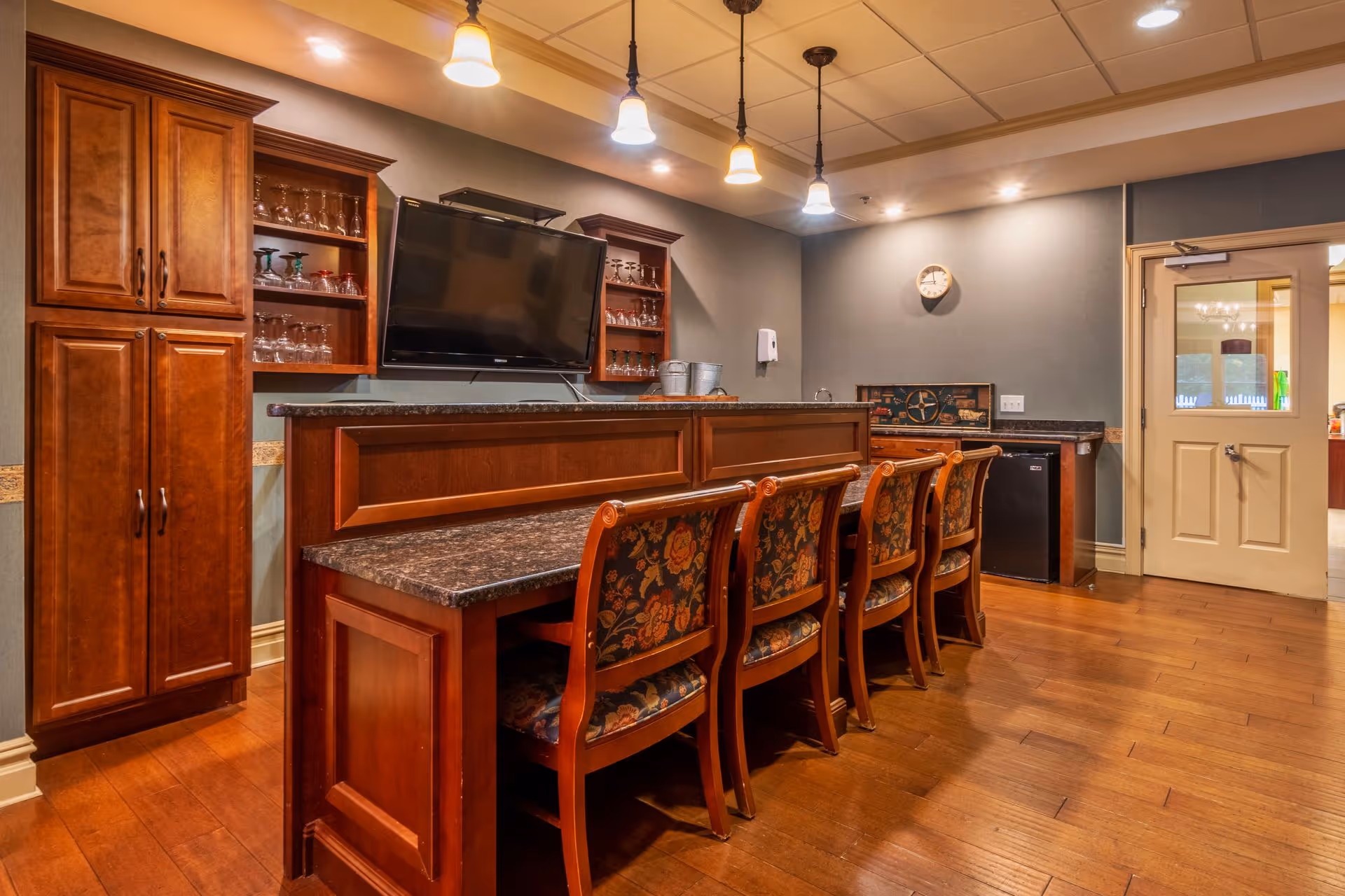 Interior view of a senior living facility's bar area with a long counter and four wooden chairs with floral upholstery. Behind the counter are wooden cabinets and shelves holding glassware, a mounted flat-screen TV, and a small refrigerator. The room has warm lighting with pendant lights hanging from the ceiling and a clock on the wall near a door with glass panels.