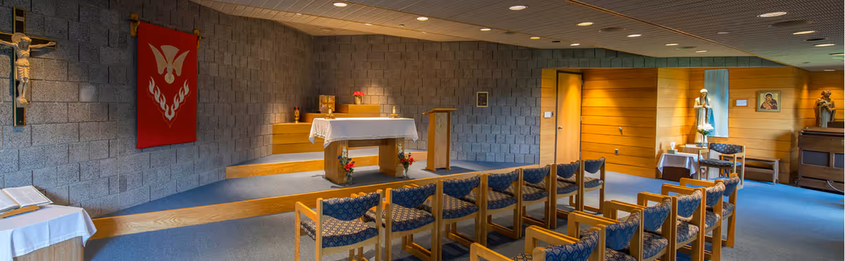 Interior view of a small chapel with rows of wooden chairs with blue patterned cushions facing an altar covered with a white cloth. The walls are made of gray cinder blocks and wood paneling. Religious symbols including a crucifix, a red banner with a dove and flames, and statues are visible around the room.